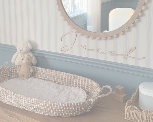 Nom en bois personnalisé Jacob en lettres attachées sous un miroir rond, au-dessus d’un panier de bébé en osier dans une chambre bleue